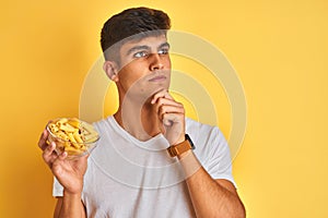 Young indian man holding bowl with dry pasta standing over isolated yellow background serious face thinking about question, very