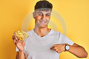 Young indian man holding bowl with dry pasta standing over isolated yellow background with surprise face pointing finger to