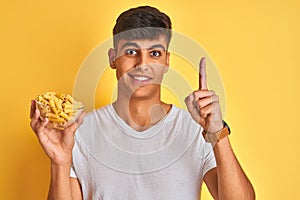 Young indian man holding bowl with dry pasta standing over isolated yellow background surprised with an idea or question pointing