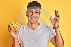 Young indian man holding bowl with dry pasta standing over isolated yellow background very happy and excited, winner expression
