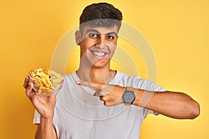 Young indian man holding bowl with dry pasta standing over isolated yellow background very happy pointing with hand and finger