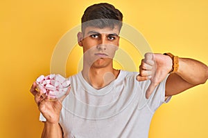 Young indian man holding bowl with marshmallows over isolated yellow background with angry face, negative sign showing dislike