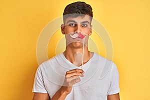 Young indian man holding fanny mustache standing over isolated yellow background with a confident expression on smart face