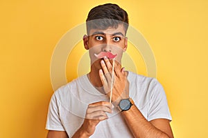 Young indian man holding fanny mustache standing over isolated yellow background cover mouth with hand shocked with shame for