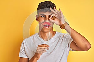 Young indian man holding fanny mustache standing over isolated yellow background with happy face smiling doing ok sign with hand