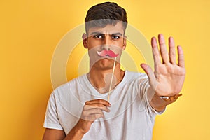 Young indian man holding fanny mustache standing over isolated yellow background with open hand doing stop sign with serious and