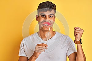 Young indian man holding fanny mustache standing over isolated yellow background screaming proud and celebrating victory and