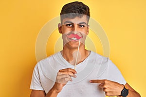 Young indian man holding fanny mustache standing over isolated yellow background with surprise face pointing finger to himself
