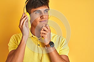 Young indian man listening to music using headphones over isolated yellow background serious face thinking about question, very