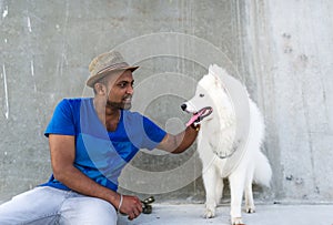 The young indian man looks at a white big dog and keeps a hand on the dog`s head.