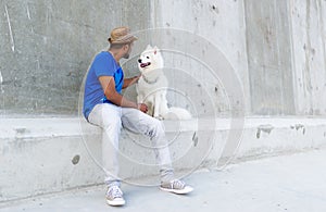 The young indian man looks at a white big dog and keeps a hand on the dog`s head. Good space on wall.