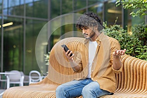 A young Indian man sits on a bench outside and looks worriedly at the phone screen, throws up his hands in frustration