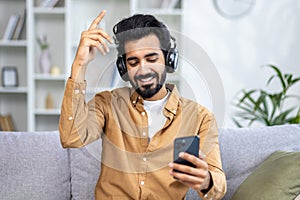 A young Indian man sits at home on the sofa with headphones, listens to music from the phone, closes his eyes, dances