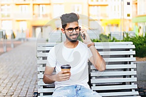 Young indian man sitting in the bench with coffee using phone in urban space