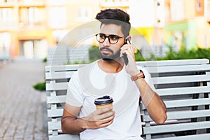 Young indian man sitting in the bench with coffee using phone in urban space