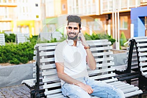 Young freelancer indian man sitting on bench, talking by phone, looking at camera. Handsome man in glasses wearing in white