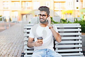 Young freelancer indian man sitting on bench, talking by phone, looking at camera. Handsome man in glasses wearing in white