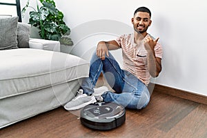 Young indian man sitting at home by vacuum robot smiling with happy face looking and pointing to the side with thumb up