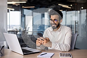 A young Indian man is sitting in an office at a desk and smilingly using a mobile phone photo