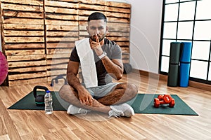 Young indian man sitting on training mat at the gym asking to be quiet with finger on lips