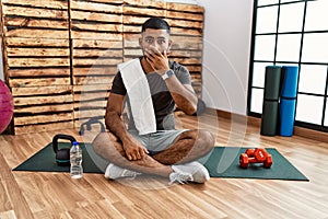 Young indian man sitting on training mat at the gym shocked covering mouth with hands for mistake