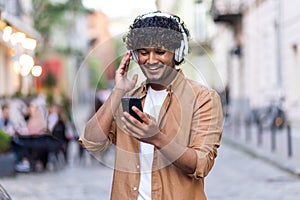 Young Indian man standing on city street wearing headphones and phone and enjoying music, smiling