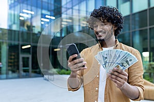A young Indian man is standing outside a skyscraper, an office building, and is holding cash money in his hands. Smiling