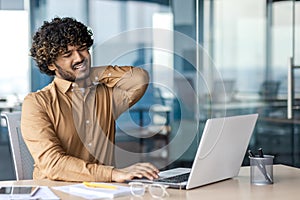 A young Indian man is suffering from severe neck pain. Tired man sits at the desk in the office and massages his hand