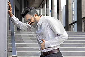 Young Indian man suffering from stomach pain, holding hands to abdomen, standing outside office building, having panic