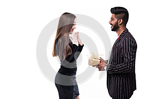 Young indian man in suit amazes his pretty asian girlfriendin dress with present wrapped in red paper, isolated on white