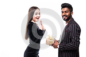 Young indian man in suit amazes his pretty asian girlfriendin dress with present wrapped in red paper, isolated on white