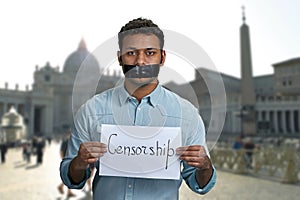 Young indian man with taped mouth holding paper card with handwritten inscription Censorship.