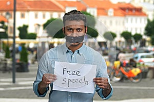 Young indian man with taped mouth holding paper card with handwritten inscription Free speech.