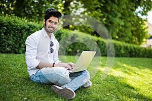 Young indian man using and typing laptop computer in summer grass in the street