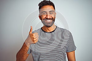 Young indian man wearing black striped t-shirt standing over isolated white background doing happy thumbs up gesture with hand