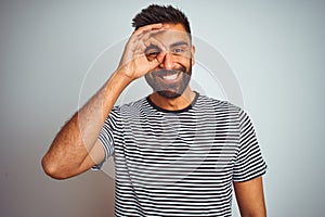 Young indian man wearing black striped t-shirt standing over isolated white background doing ok gesture with hand smiling, eye