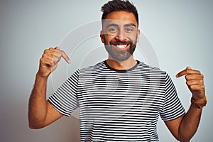 Young indian man wearing black striped t-shirt standing over isolated white background looking confident with smile on face,