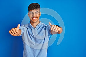 Young indian man wearing casual polo standing over isolated blue background approving doing positive gesture with hand, thumbs up