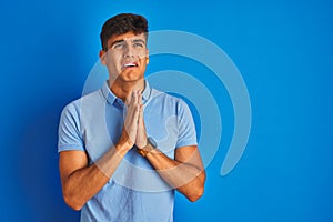 Young indian man wearing casual polo standing over isolated blue background begging and praying with hands together with hope