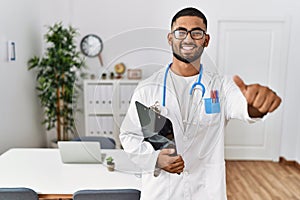 Young indian man wearing doctor uniform and stethoscope approving doing positive gesture with hand, thumbs up smiling and happy