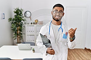 Young indian man wearing doctor uniform and stethoscope pointing to the back behind with hand and thumbs up, smiling confident