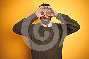 Young indian man wearing green sweater and shirt standing over isolated yellow background doing ok gesture like binoculars