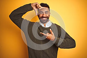 Young indian man wearing green sweater and shirt standing over isolated yellow background smiling making frame with hands and