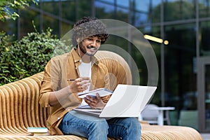 A young Indian man wearing headphones is sitting on a street bench, holding a laptop on his lap and making notes in a