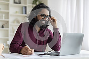 Young indian man wearing headset using laptop and smiling while taking notes