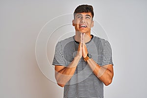Young indian man wearing navy striped t-shirt standing over isolated white background begging and praying with hands together with
