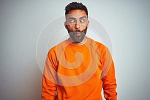 Young indian man wearing orange sweater over  white background making fish face with lips, crazy and comical gesture