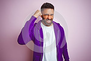 Young indian man wearing purple sweatshirt standing over isolated pink background angry and mad raising fist frustrated and