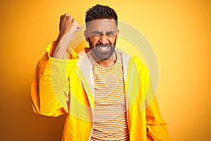 Young indian man wearing raincoat standing over isolated yellow background angry and mad raising fist frustrated and furious while