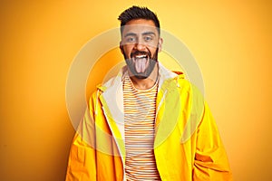 Young indian man wearing raincoat standing over isolated yellow background sticking tongue out happy with funny expression